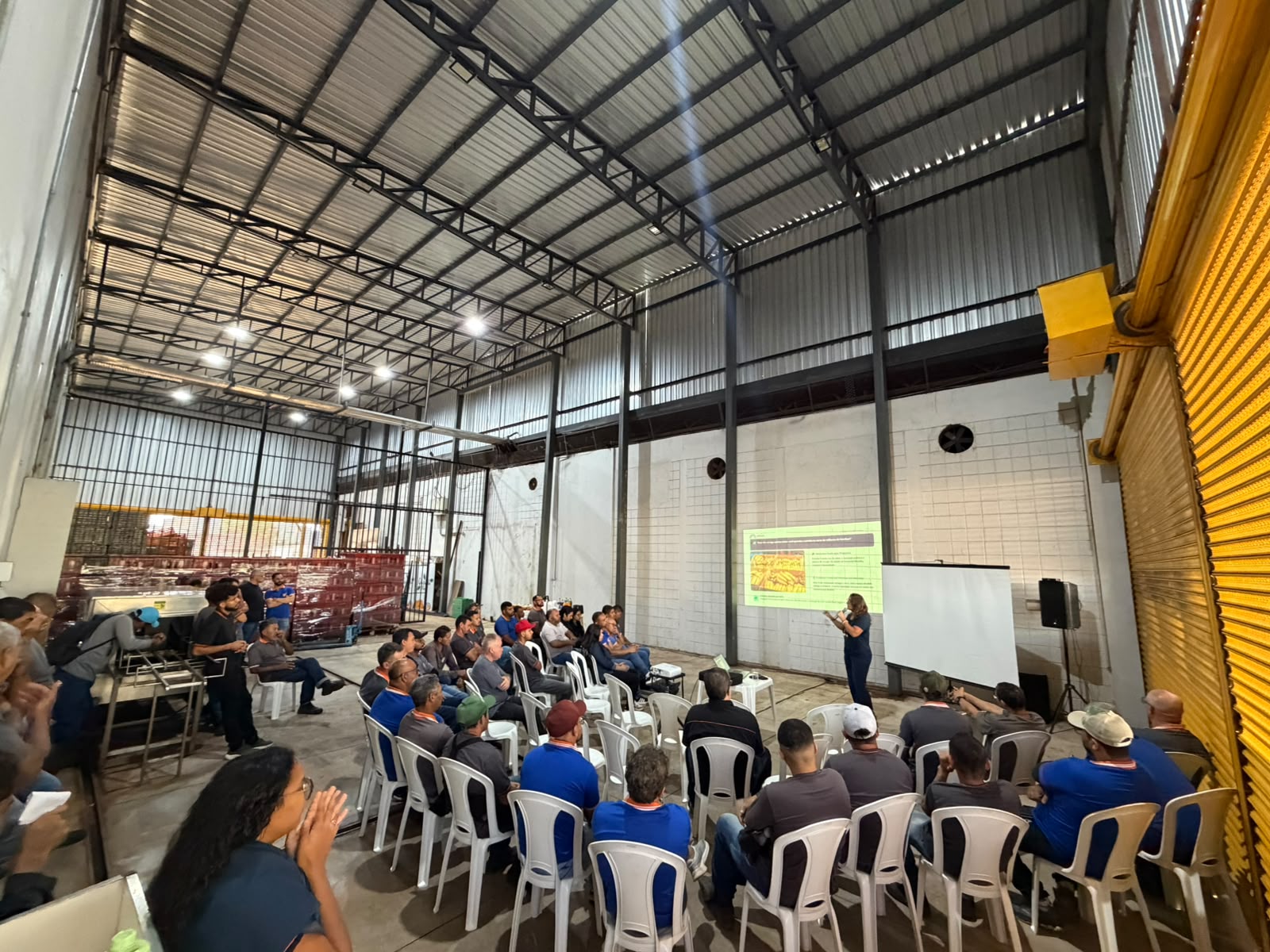 Vista ampla do galpão da Comercial Mendes durante a palestra, com a equipe sentada e o slide projetado na parede