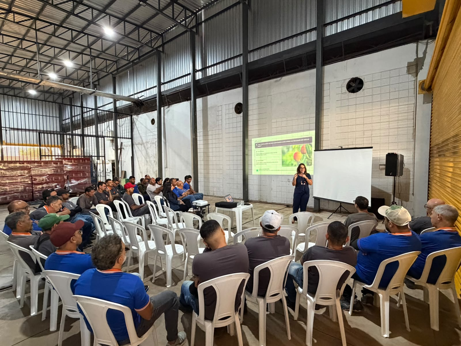 Psicóloga da Arcus Psicologia apresentando a palestra para a equipe da Comercial Mendes sentada em cadeiras brancas no galpão